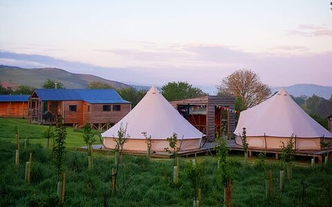 A photograph of two bell tents with a log cabin and hills in the background
