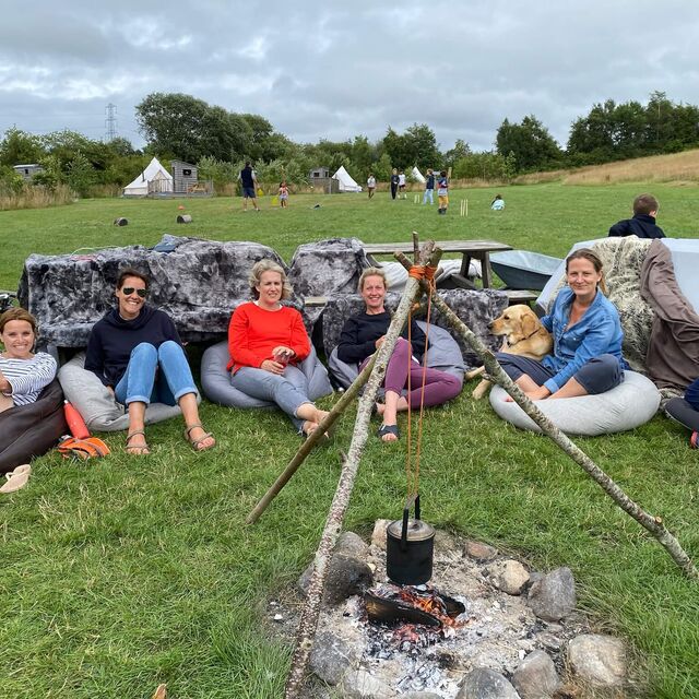 A photo of a group of friends sitting around a campfire with children playing cricket in the background