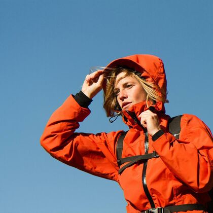 A young woman in red hiking jacket raises her hood, no other details can be seen except a clear blue sky.