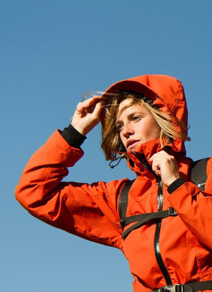A young woman in red hiking jacket raises her hood, no other details can be seen except a clear blue sky.