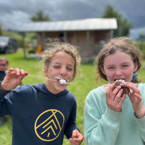 A photograph of two girls eating marshmallows with a cabin in the background