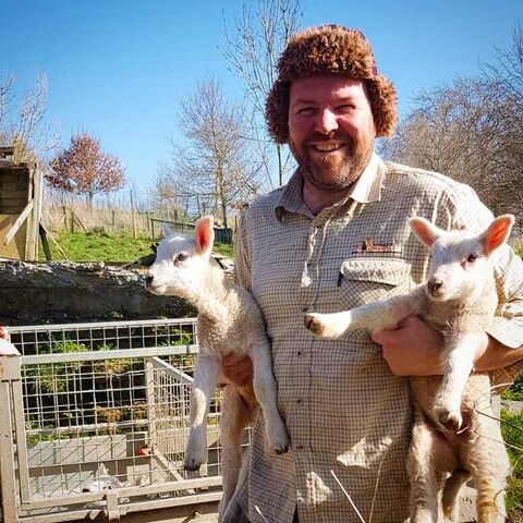 A photo of a man in a hat holding a small lamb under each arm