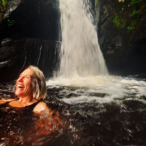 A photograph of a woman in a waterfall with a look of pure joy and happiness on her face.